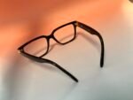 Close-up of black eyeglasses with a gold hinge, Versace Made in Italy printed on the temple on a peach gradient background.
