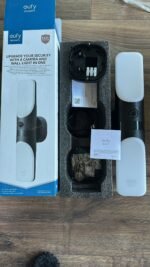 Boxed Eufy Security wall light camera kit with components laid out: light panel, camera unit, wires and manuals visible on a wooden floor.