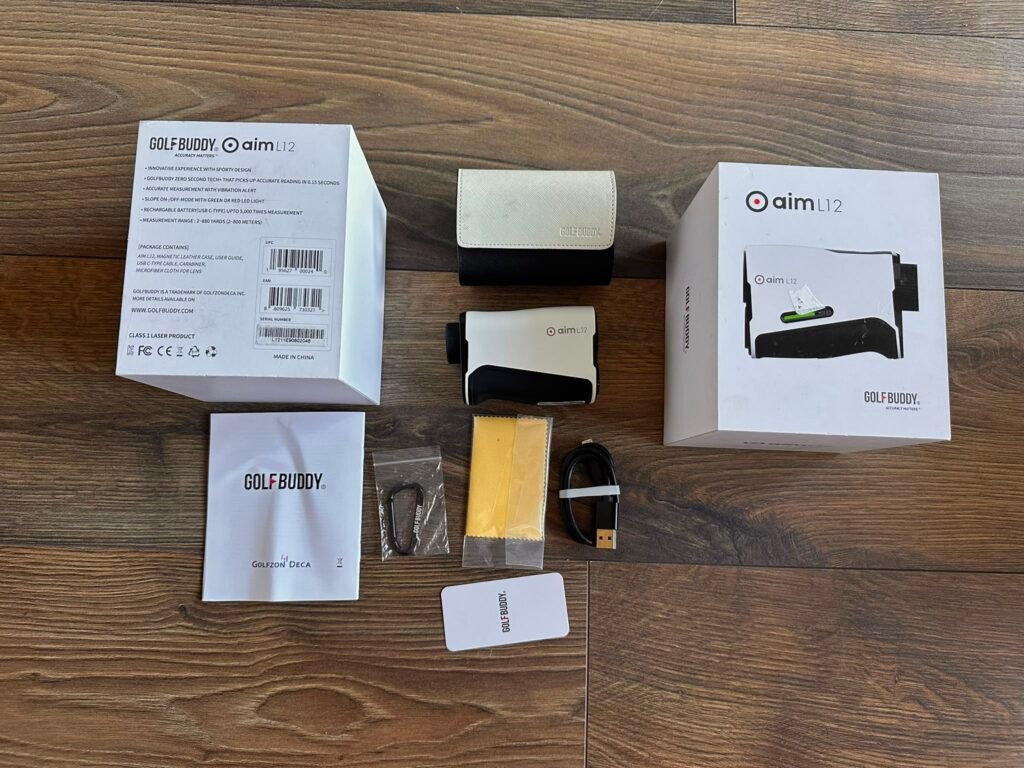 Unopened GolfBuddy AIM L12 package with white box on left, mid-height device and accessories laid out on a wooden floor including a beige leather case and a white box labeled 'aim L12' on the right.