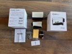 Unopened GolfBuddy AIM L12 package with white box on left, mid-height device and accessories laid out on a wooden floor including a beige leather case and a white box labeled 'aim L12' on the right.