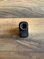 Close-up of a small black camera or action camera resting on a wooden surface, showing its circular lens and a red status light.