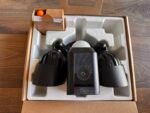 Top-down view of a black VR headset in its foam packaging inside a brown box on a wooden floor, with a small separate box containing an orange ball.