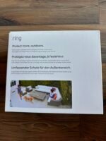 White Ring product box with the slogan 'Protect more, outdoors' and a photo of an outdoor patio with seating and a dog visible at the bottom left corner.