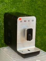 Smeg espresso machine with a brushed metal front and black body on a counter, green hedge in the background