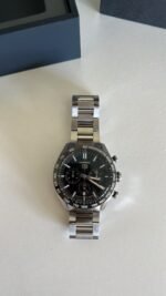 Stainless steel chronograph watch with black dial and tachymeter bezel on a white surface, blue box in background.