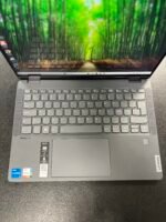 Open Lenovo laptop on a dark desk displaying a vibrant forest wallpaper across the screen and keyboard area visible below.